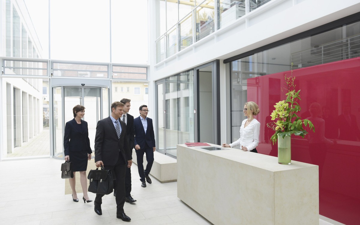 5 business professionals dessed in dark formal clothing arrive in a glazed reception area and are greeted by a receptionist wearing white. There is a desk with flowers at one end and the is a crimson red screen behind plate glass. The floor is white.