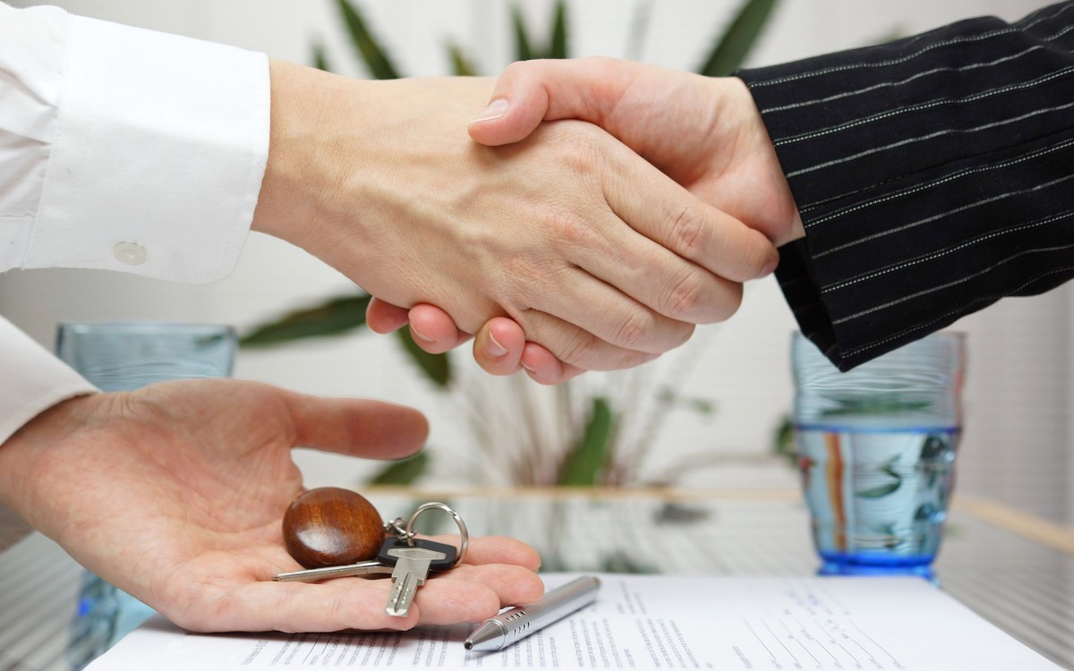 Three hands are visible belonging to two people. On the left we see two hands of 1 person, one is in a hand shake clasp with another person. The second hand of the first person is holding a key with an attractive dark wood keyring. 