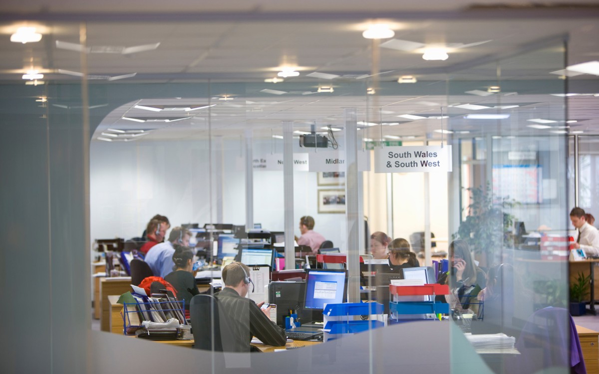 A busy office space as viewed through plate glass, people working in front of computer screens and wearing headsets