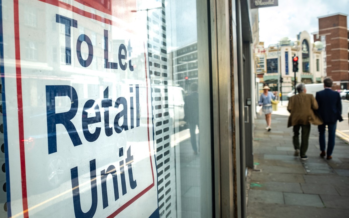 To let retail unit sign in window, urban setting, people are walking down the pavement, reflected in the window along with cars. The letters are blue surrounded by a red border.